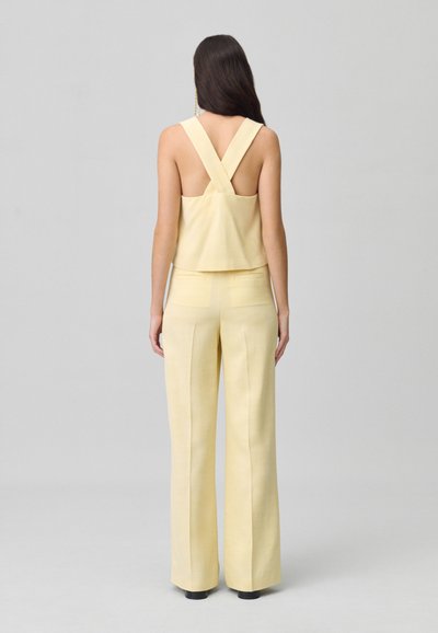 Top corto giallo con spalline incrociate abbinato a pantaloni a vita alta e gamba larga, entrambi con una texture liscia e un design minimalista.