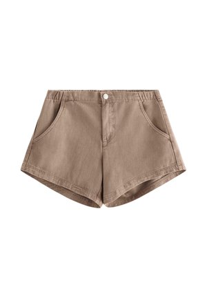 Unge person stående, iført lysblå mønstret bluse med blonderærmer, brune shorts og beige snørebåndssko på ensfarvet baggrund.