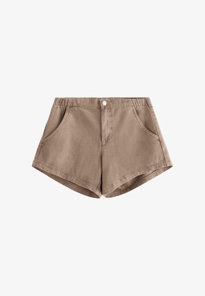 Unge person stående, iført lysblå mønstret bluse med blonderærmer, brune shorts og beige snørebåndssko på ensfarvet baggrund.