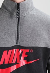 Šedá mikina na zip do půlky se spodní černou částí a velkým červeným logem "NIKE" přes hruď. Měkká látka s hladkou texturou.