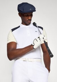 Biała koszulka polo z granatowymi i jasnobeżowymi akcentami, białym paskiem i białą rękawiczką golfową. Granatowy płaski kapelusz. Trzyma kij golfowy.