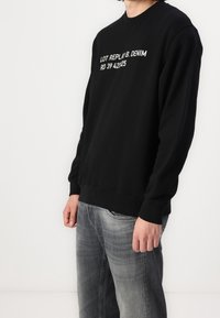 Felpa nera con collo rotondo e maniche lunghe. Presenta del testo bianco sul davanti. Abbinata a jeans grigi chiari.