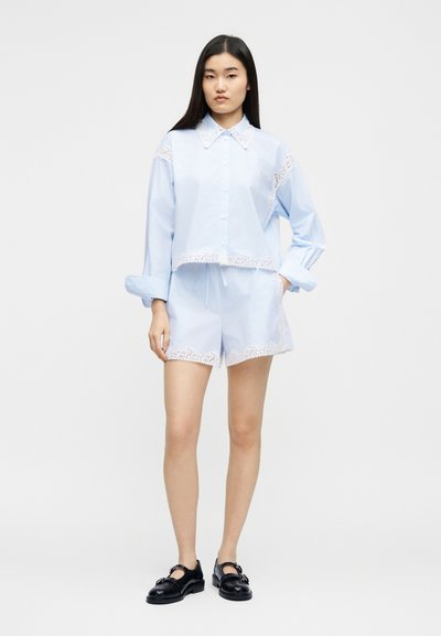 Νεαρή γυναίκα με γαλάζιο πουκάμισο με κουμπιά και κοντό παντελόνι με λευκή δαντέλα στο τελείωμα, συνδυασμένα με μαύρα παπούτσια Mary Jane, στέκεται μπροστά σε λευκό φόντο.