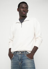 Balta jebs ar rāvējslēdzēju, augstu apkakli, brīvs griezums un ribotas piedurknes, apvienota ar zilām nodilušām džinsiem un pelēku T-kreklu zem tā.