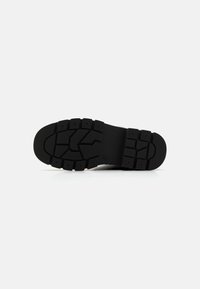 Semelle de chaussure noire présentant un design en caoutchouc texturé, avec des crampons proéminents pour l'adhérence. Bords lisses et zone de cheville plate en contraste.