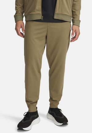 Mann trägt olivgrüne Jogginghosen, schwarze Sneaker mit weißen Sohlen und eine olivgrüne Jacke über einem schwarzen Hemd.