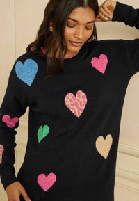 Fekete kötött pulóver, színes szívmintákkal kék, rózsaszín, zöld és leopárdmintás változatban. Bordázott mandzsetták és rózsaszín nyakkivágás.