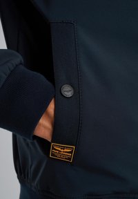 Marineblaues Jackett mit gerippten Bündchen, glatter Textur, Seitentasche und einem runden Logoknopf. Verfügt über ein rechteckiges Markenlabel in Orange.