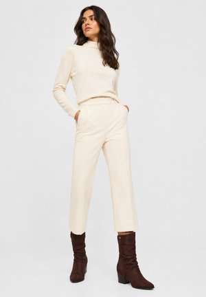 Femme aux longs cheveux foncés portant un pull à col roulé crème, un pantalon large assorti et des bottines en daim brun foncé, debout les mains dans les poches.