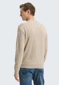 Beige gebreide trui met lange mouwen, ronde hals en geribbelde zoom, gecombineerd met een blauwe spijkerbroek. De stof lijkt zacht met een gladde textuur.