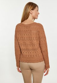 Maglione marrone a maglia con motivi ad uncinetto, scollo rotondo, polsini e orlo a coste. Indossato con pantaloni beige, che mostrano un design casual.