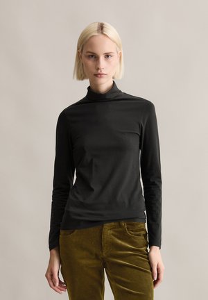 Zwarte langemouwen turtleneck shirt gemaakt van gladde stof, gedragen met olijfgroen corduroy broek. Eenvoudig, aansluitend ontwerp met een hoge kraag.