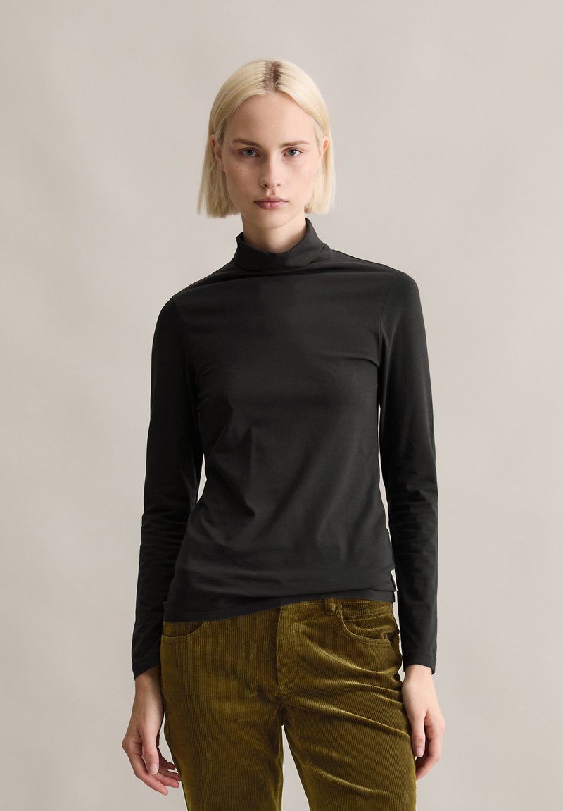 Zwarte langemouwen turtleneck shirt gemaakt van gladde stof, gedragen met olijfgroen corduroy broek. Eenvoudig, aansluitend ontwerp met een hoge kraag.
