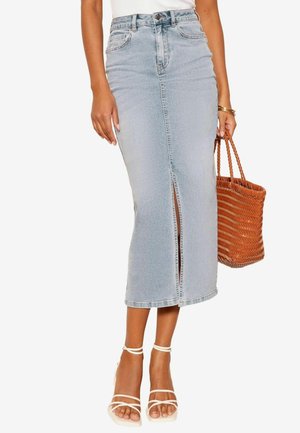 Femme portant une jupe midi en jean bleu clair taille haute avec une fente à l'avant, des talons blancs à brides, des bracelets dorés et tenant un sac à main tissé marron.