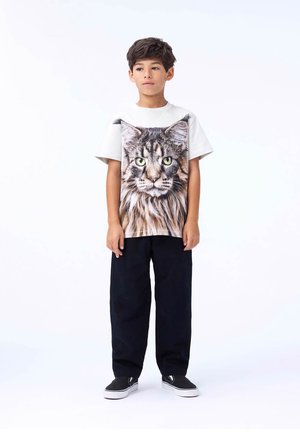 T-shirt blanc avec un grand motif de chat sur le devant, portant un pantalon noir ample et des baskets noires, se tenant devant un fond blanc.