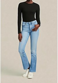 Personne portant un haut noir à manches longues, un jean bootcut taille haute bleu clair, et des chaussures blanches à lacets sur un fond neutre.