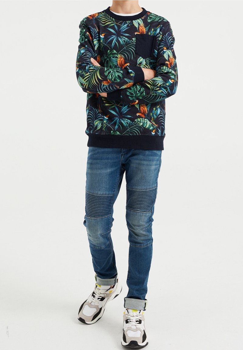 Sweat-shirt bleu marine avec imprimé de feuilles tropicales et d'oiseaux, poignets côtelés, col rond ; associé à un jean skinny bleu clair avec accents côtelés aux genoux et baskets.