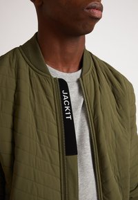Giacca bomber trapuntata verde oliva con polsini a costine; presenta un'etichetta nera con la scritta "JACK1T" sulla cerniera; indossata sopra una maglietta grigia.
