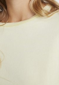 Hellgelbes T-Shirt mit einer weichen, glatten Textur und einem runden Ausschnitt. Verfügt über ein einfaches und klares Design, teilweise sichtbarer Hintergrund.