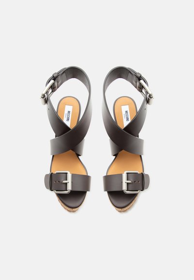 Καφέ δερμάτινα σανδάλια με φαρδιές σταυρωτές λουρίδες, ασημένιες αγκράφες στους αστραγάλους και στα δάχτυλα, και ταμπά επενδυμένες εσωτερικές σόλες με την ετικέτα Moschino.