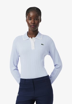 Donna con capelli intrecciati che indossa una polo a maniche lunghe a coste di colore azzurro chiaro e pantaloni blu scuro, in piedi con le mani dietro la schiena.