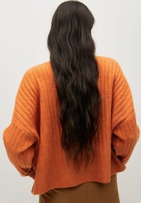 Pull en maille côtelée orange avec un design court et des manches larges, présentant une surface texturée. Porté sur un bas marron.