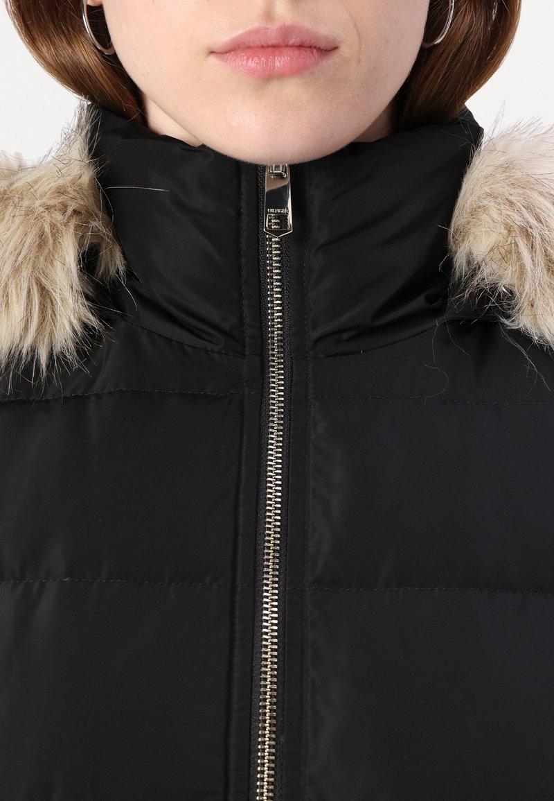 Close-up van een persoon die een zwarte pufferjack met een bontgevoerde capuchon draagt en een zilveren rits die tot aan de nek is dichtgeritst.