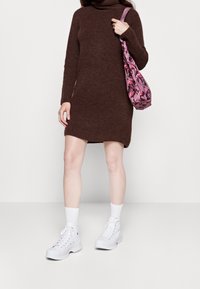 Abito a collo alto in maglia marrone con aperture laterali, abbinato a sneakers bianche alte e una borsa a tracolla rosa con motivo.