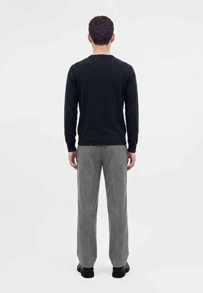 Homme debout de dos, portant une chemise noire à manches longues, un pantalon gris et des chaussures noires sur un fond blanc uni.
