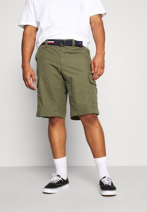 Homme portant un short cargo vert olive avec des poches latérales, une ceinture blanche, des chaussettes blanches montantes, des baskets noires et un t-shirt blanc, debout contre un mur blanc.