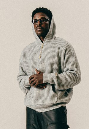Man with glasses wearing a gray knit hoodie and black pants, standing with hands clasped against a plain background.