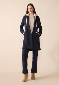 Cappotto blu navy con cappuccio foderato in beige, tasche frontali e chiusura con bottoni, abbinato a pantaloni blu scuro e stivaletti beige chiaro alla caviglia.