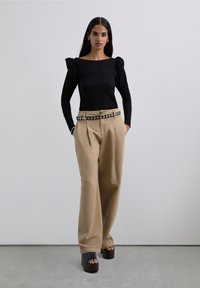 Woman standing with hands in pockets wearing black puff-sleeve top, beige wide-leg trousers with studded belt, and black platform sandals.