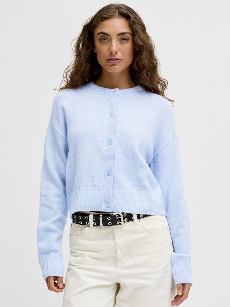 Jauna moteris su banguotais rudais plaukais, dėvinti šviesiai mėlyną megztinį su sagomis, baltas aukštakulnes kelnes ir juodą diržą su metalinėmis apkaustais, stovinti prieš baltą foną.