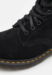 Black suede ankle boot with black laces, metal eyelets, and a yellow stitched sole. Features a smooth texture and structured design.
