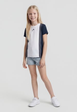 Šypsosi mergina su ilgais šviesiais plaukais, vilkinti trumpomis rankovėmis su spalvų blokais marškinėliais, džinsiniais šortais ir baltais sportbačiais, stovinti pilkame fone.