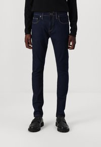 Sötétkék, slim fit farmernadrág, világos varrással, elülső zsebekkel és gombos záródással. Fekete cipővel viselve.