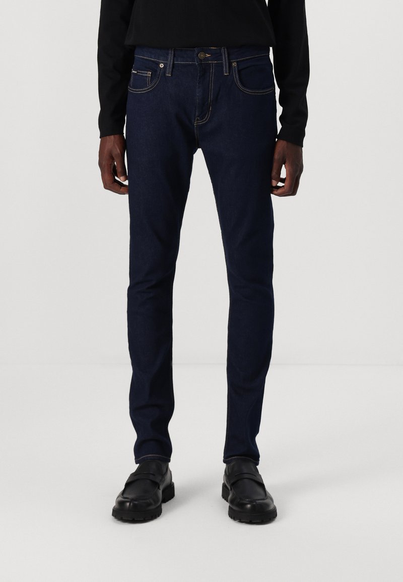 Sötétkék, slim fit farmernadrág, világos varrással, elülső zsebekkel és gombos záródással. Fekete cipővel viselve.