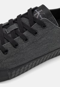 Šedá textilní teniska s černými tkaničkami a oky. Texturovaný svršek, gumová podrážka s výraznými drážkami, s logem na jazyku.