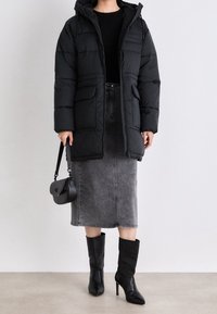 Kobieta ubrana w czarną pikowaną kurtkę, szarą midi spódnicę dżinsową, czarne botki na wysokim obcasie i trzymająca małą czarną torebkę.