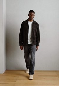 Black corduroy jacket over a white t-shirt, paired with gray jeans and beige sneakers; standing on wooden floor against a neutral backdrop.