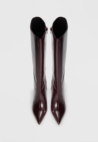 Une paire de bottes en cuir marron foncé, à bout pointu et hauteur élevée, avec des fermetures éclair latérales, se tenant verticalement sur un fond clair et uni.