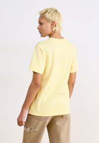 T-shirt en coton jaune à manches courtes et à encolure ronde, vue de dos. Assorti à un pantalon beige clair avec un ourlet effiloché.