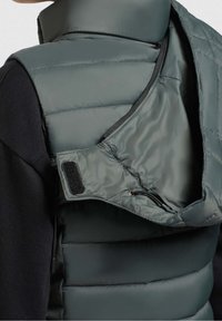 Gilet imbottito verde scuro con collo alto. Presenta una chiusura con zip e dettagli in Velcro. L'interno mostra una trama liscia e lucida.
