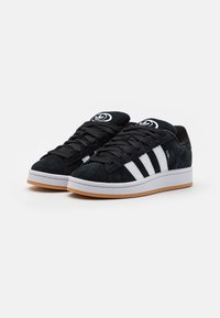 Baskets en daim noires avec un design à trois rayures blanches et un logo, dotées de lacets noirs et d'une semelle en caoutchouc gomme.