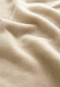 Beige neulottu kangas, jossa on sileä pinta, esittelee hienovaraisia kudontavaihteluita ja pehmeitä ryppyjä, mikä viittaa pehmeyteen ja muokattavuuteen.