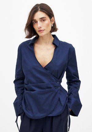 Navy blue wrap shirt in a smooth fabric with a deep V-neck, long sleeves, side ties, and button closures for adjustable fit.