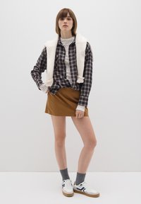 Modella che indossa una camicia quadrettata blu navy e bianca, sovrapposta a un maglione crema, abbinata a una mini gonna marrone e sneakers bianche con accenti blu navy.