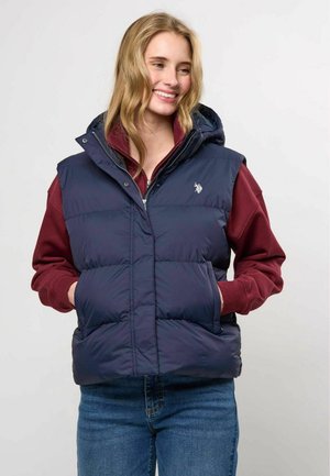 Jeune femme souriante portant une doudoune bleu marine par-dessus un sweat à capuche bordeaux, les mains dans les poches de la doudoune et un jean bleu sur un fond clair.