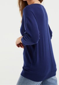 Tmarinová modrá pletená mikina se třičtvrtinovými rukávy, žebrovanými manžetami a spodním lemem, s centrálním švem na zadní straně. Měkký, lehký materiál.